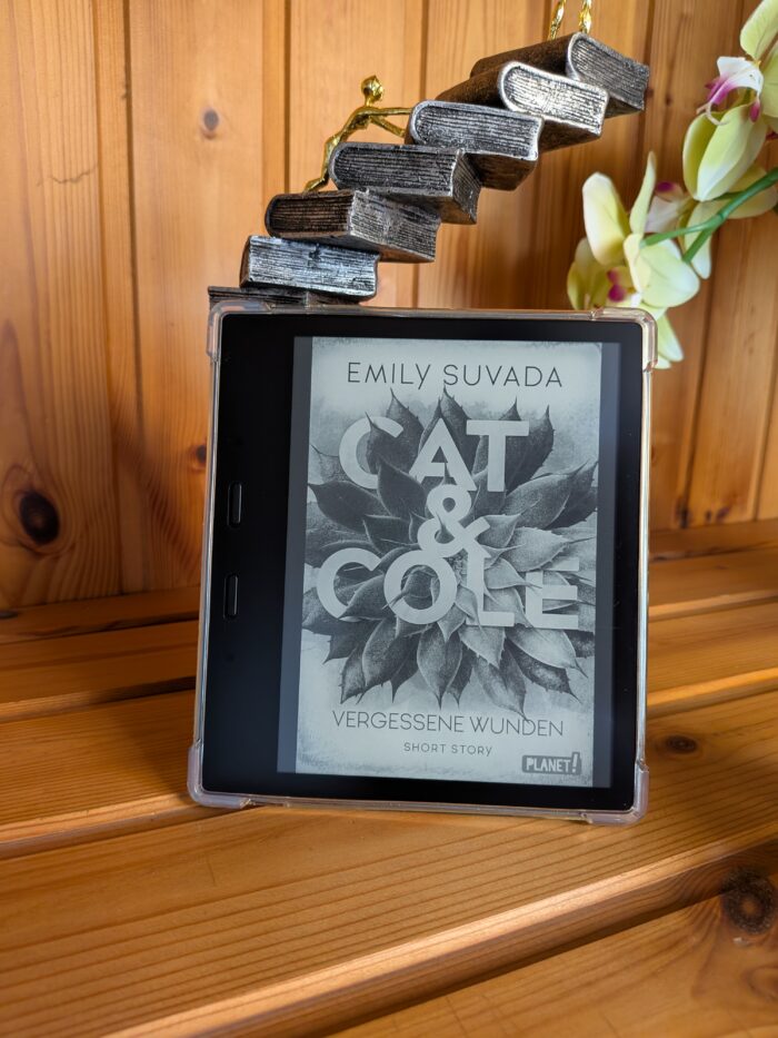 Emily Suvada: Cat & Cole - Vergessene Wunden. Im Vordergrund steht ein Kindle mit dem Cover in schwarz-weiß. Eigentlich ist es ein weißes Cover mit einer orangenen Blume, der integrierte Schriftzug ist weiß. Der Hintergrund sind Holzdielen und eine Bücherturmfigur und eine Orchideenblüte.