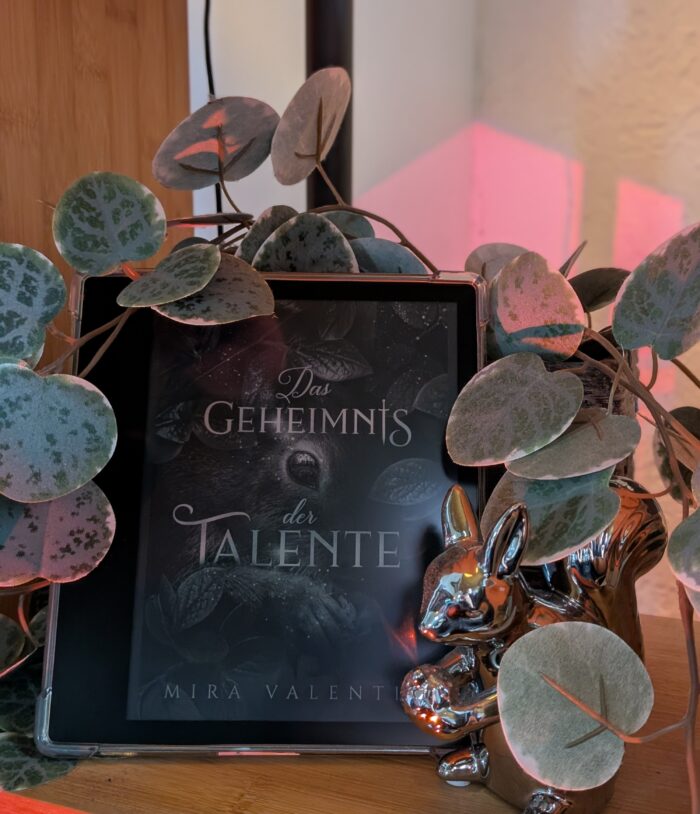 Mira Valentin - Das Geheimnis der Talente. Ein Kindle mit dem Cover des Buches und eine silberne Eichhörnchenfigur werden umrahmt von Blättern. Auf dem Cober ist ein Eichhörnchen in Blättern versteckt mit dem Titel im Vordergrund. Eigentlich ist das Eichhörnchen orange mit grünen Augen und grünen Blättern, doch das Kindle zeigt es in schwarz-weiß.