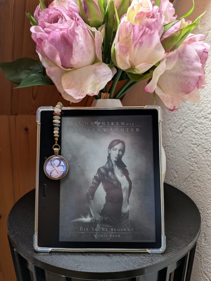 Nicole Böhm: Die Chroniken der Seelenwächter - Die Suche beginnt. Auf dem Cover ist die Hauptprotagonistin Jess mit einem Dolch zu sehen. Der Kindle steht vor einer Vase mit rosa Blumen auf einem schwarzen Tisch. An dem Kindle hängt ein Lesezeichen mit dem Symbol der Seelenwächter, 2 ineinander verschlungene Unendlichkeitszeichen (Achten).