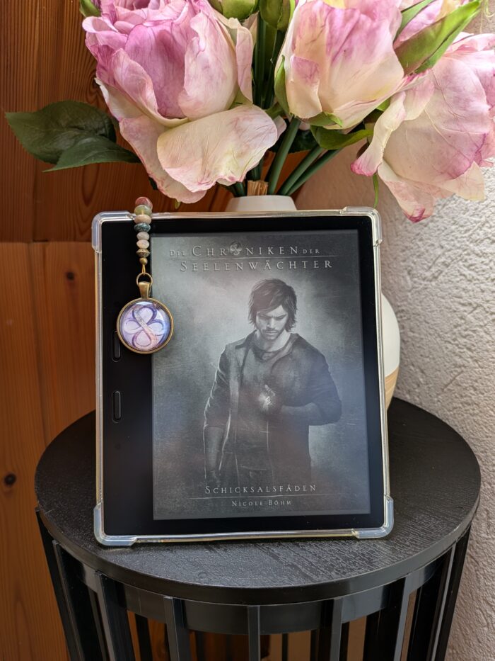 Nicole Böhm: Die Chroniken der Seelenwächter - Schicksalsfäden. Auf dem Cover sieht man den Hauptprotagonisten Jaydee. Der Kindle steht auf einem Tisch, im Hintergrund sieht man eine Vase mit rosa Blumen. Am Kindle hängt das Symbol der Seelenwächter, zwei ineinander verschlungene Unendlichkeitszeichen (Achten).