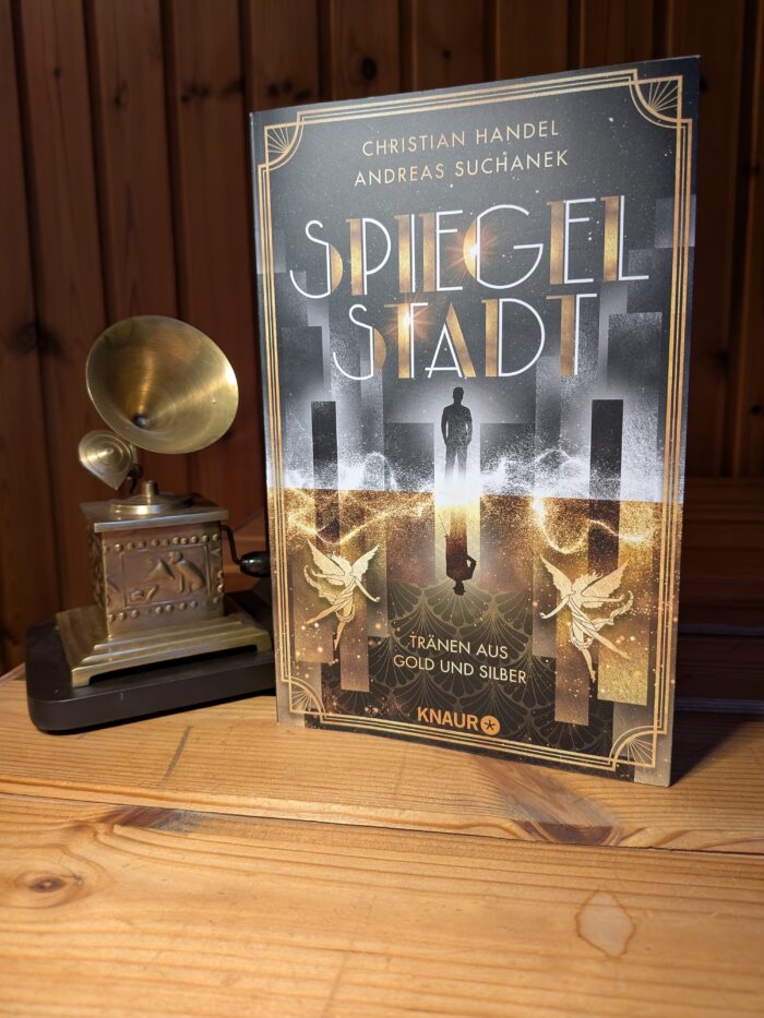 Christian Handel + Andreas Suchanek: Spiegelstadt - Tränen aus Gold und Silber. Das Buch mit silber und goldenen Cover ist zu sehen, darauf spiegelt sich ein Mann. Auf der oberen silbernen Seite sieht man den Mann in der Jetzt-Zeit und auf der unteren, goldenen Seite sieht man ihn im Stile der 20iger Jahre angezogen. Außerdem sieht man zwei goldene Feen. Links daneben steht ein Grammophon, als Deko-Element.