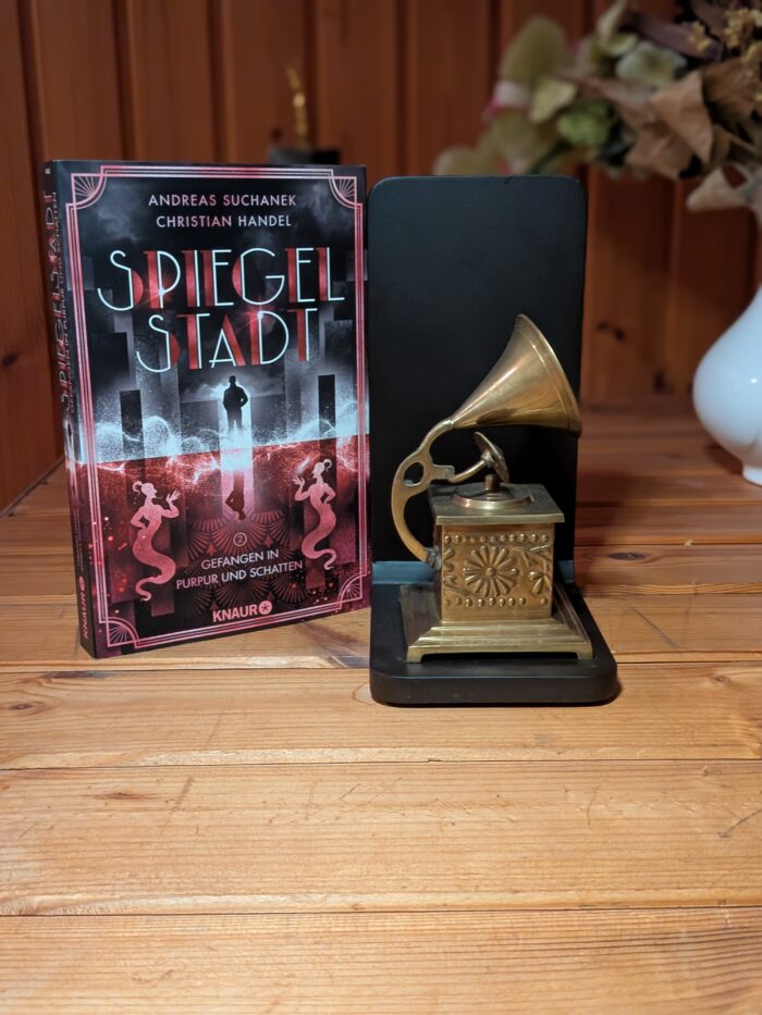 Andreas Suchanek + Christian Handel: Spiegelstadt - Gefangen in Purpur und Schatten. Links ist das Taschenbuch mit silbernen und roten Hintergrund. Im silbernen Teil ist ein Mann aus der Jetztzeit und im roten Teil ein Mann in den zwanziger Jahren, außerdem zwei Dschinn. Daneben steht ein Grammophon in Gold, als Dekoelement.