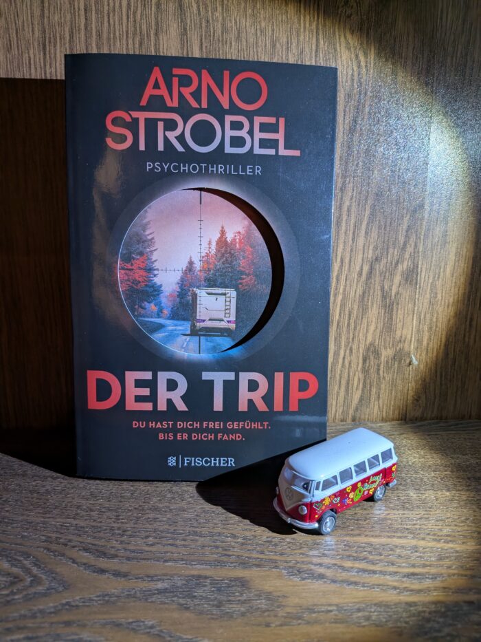 Arno Strobel: Der Trip - Du hast dich frei gefühlt. Bis er dich fand. Im Regal steht das Buch in schwarz gehalten, mit roter Schrift. Darauf zu sehen ist, ein Wohnwagen auf der Straße, darüber ist ein Fadenkreuz von einem Zielfernrohr. Neben dem Buch steht eine Miniaturausführung eines VW Bulli.