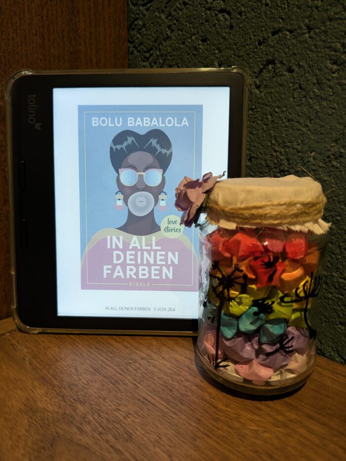 Bolu Babalola: In all deinen Farben. Ein Tolino mit dem Cover einer nigerianischen Frau mit bunter Kleidung und dem Titel in weiß geschrieben. Daneben steht ein Glas mit bunten, gebastelten Sternen.