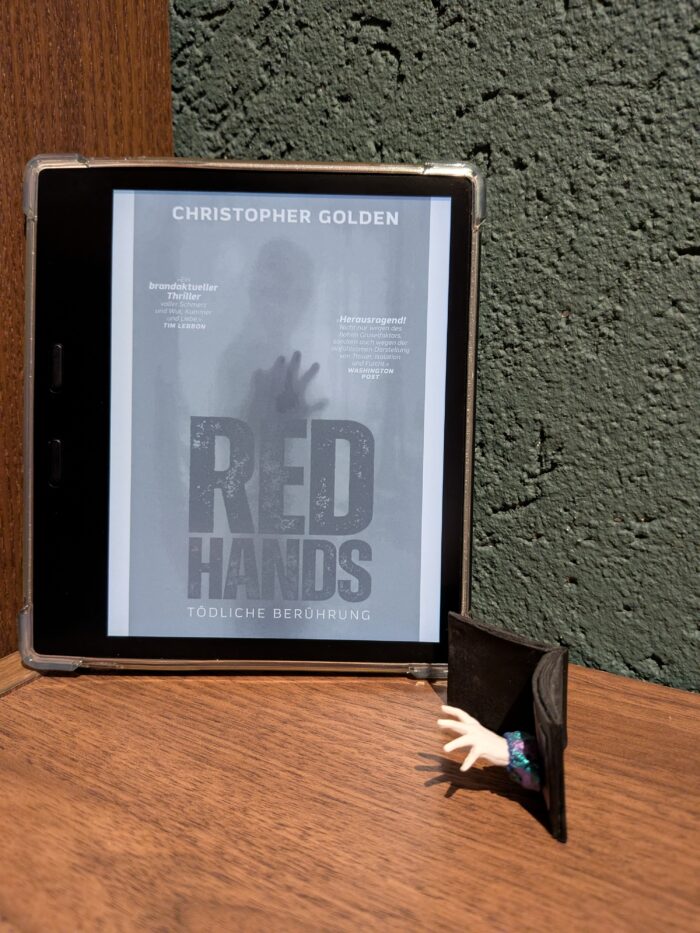 Christopher Golden: Red Hands - Tödliche Berührung. Auf einem Regal steht ein Kindle, mit dem Cover vom Buch, daneben steht eine Miniaturausgabe eines Buches, aus dem eine Hand herauskommt. Das Cover ist hellblau mit roter Schrift, im Hintergrund sieht man einen Menschen, der seine rote Hand ausstreckt.