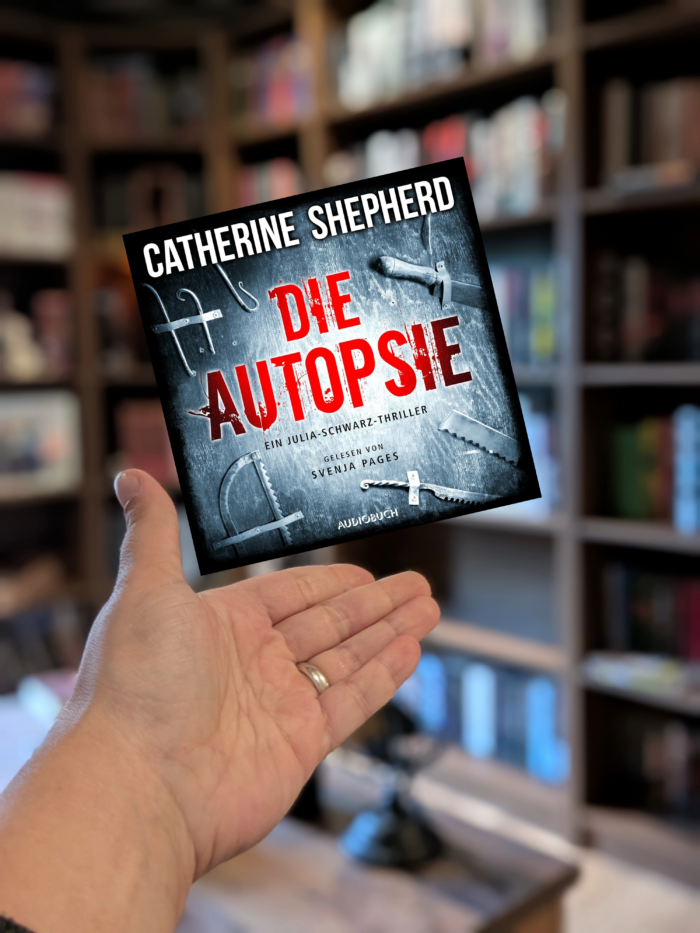 Caterine Shepherd: Die Autopsie - Ein Julia-Schwarz-Thriller. Man sieht eine Hand, darüber das Cover des Hörbuches. Das Cover ist in schwarz-weiß gehalten, mit weißer und roter Schrift. Außerdem sieht man Sägen und Haken. Im Hintergrund sieht man Bücherregale.
