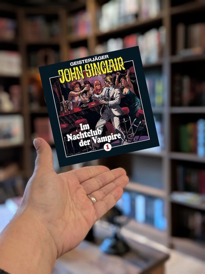 Jason Dark: Geisterjäger John Sinclair - Im Nachtclub der Vampire. Eine Hand präsentiert ein Hörbuchcover, welches in schwarz gehalten ist, mit weißer und gelber Schift. Darauf sieht man einen blonden Mann in einer Bar, umringt von Vampirinnen. Im Hintergrund sieht man verschwommen ein Bücherregal.