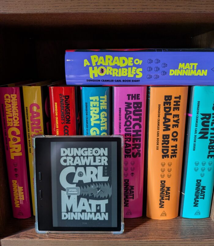Matt Dinniman: Dungeon Crawler Carl. Im Hintergrund in einem Regal sind alle englischen Ausgaben der Reihe, im Vordergrund ein Kindle mit dem schwarz-weißen Cover. 