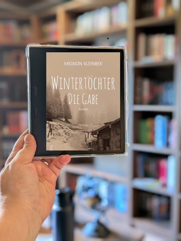 Mignon Kleinbek: Wintertöchter Trilogie. Im Vordergrund ist ein Kindle Cover des Buches, dieses ein beiges Winterbild mit weißer Schrift.
