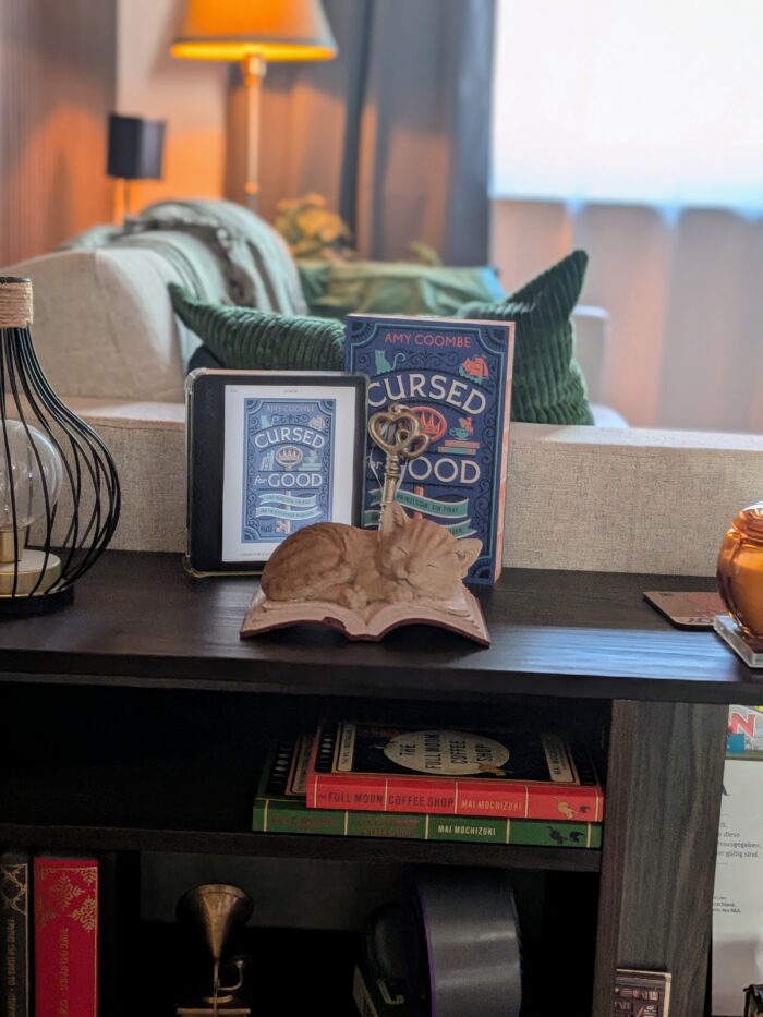 Amy Coombe: Cursed for Good - Eine Prinzessin. Ein Pirat. Und ein verfluchter Buchladen: Im Hintergrund ist ein helles Wohnzimmer, im Vordergrund ein kleines Regal mit dem Buch und einem Tolino mit dem Cover des Buches. Außerdem einen großen Schlüssel und eine Deko Katze schlafend auf einem Buch.
